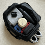 Black backpack with various items including a water bottle, shoes, and a vibit roller on a beige carpet.