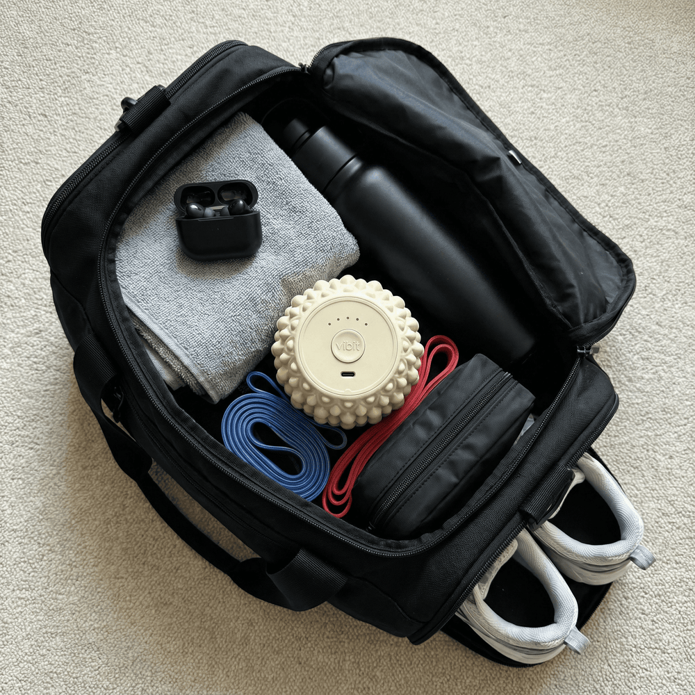 Black backpack with various items including a water bottle, shoes, and a vibit roller on a beige carpet.